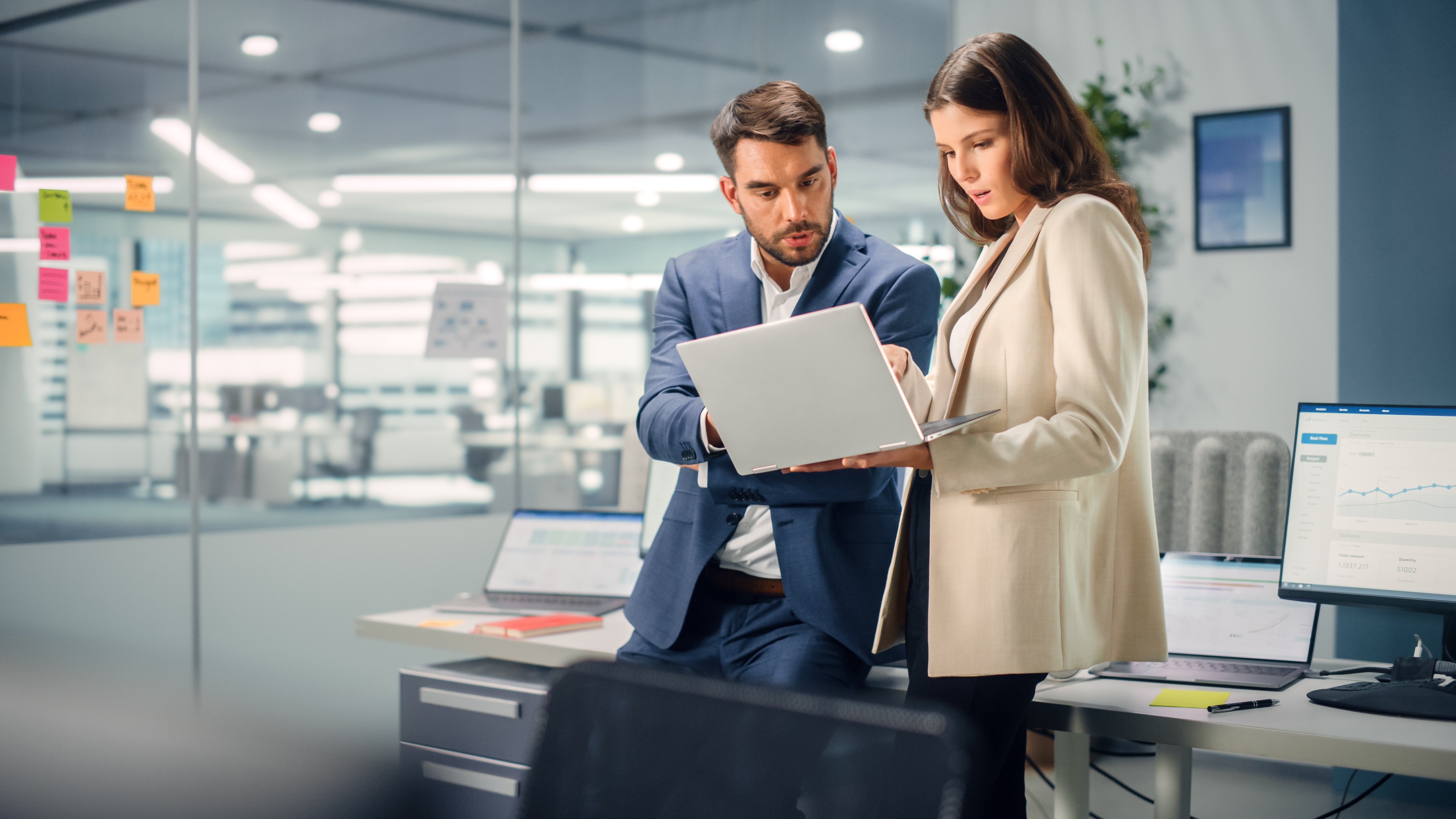 A male and female consumer loan lender looking at client portfolio performance reports on a laptop in a well-lit large office space